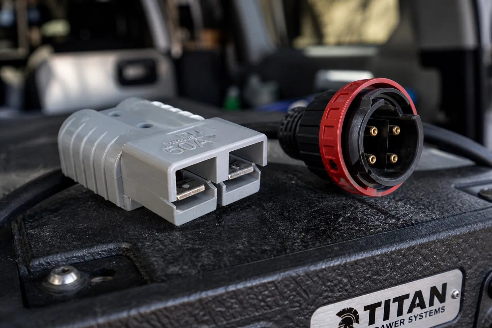A comparison of different 12V power plugs including a standard cigarette lighter plug, a Merit plug, and a grey 50A Anderson plug, displayed on an Australian 4WD workshop bench.