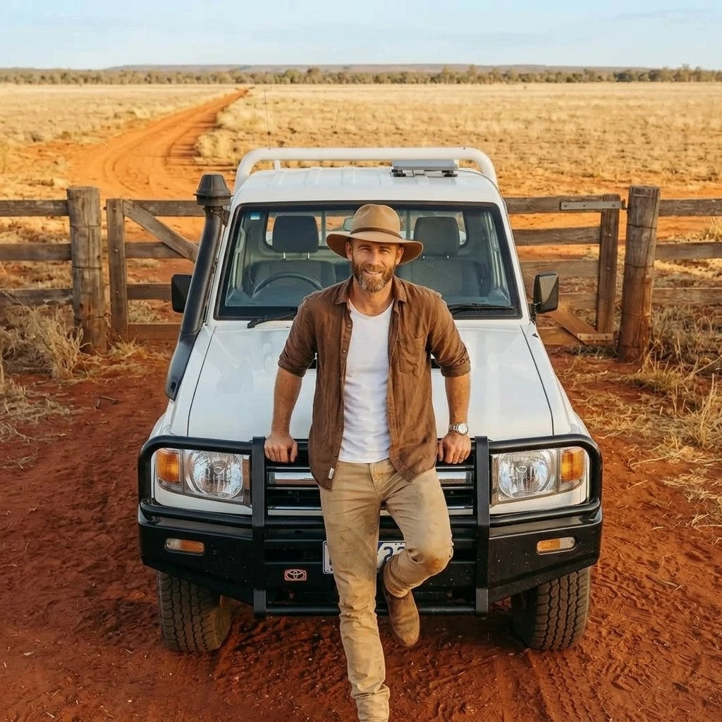 Bloke in akubra and brown shirt leaning on the bullbar of a white Toyota 70-series troopy at a station gate, open paddock and red dirt track behind, Starlink Mini satellite dish mounted flat on the troopy roof on a charcoal alloy magnetic mount.
