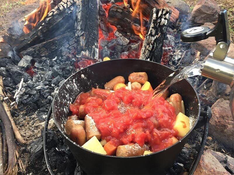 A steaming cast-iron camp oven filled with hearty chicken and vegetable stew sitting on red dirt next to a campfire.