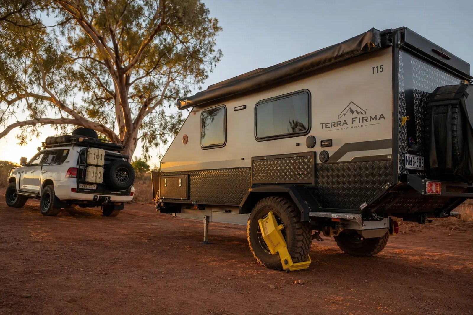 A high-end Australian off-road caravan with a yellow wheel clamp fitted at a remote red-dirt campsite at sunset.