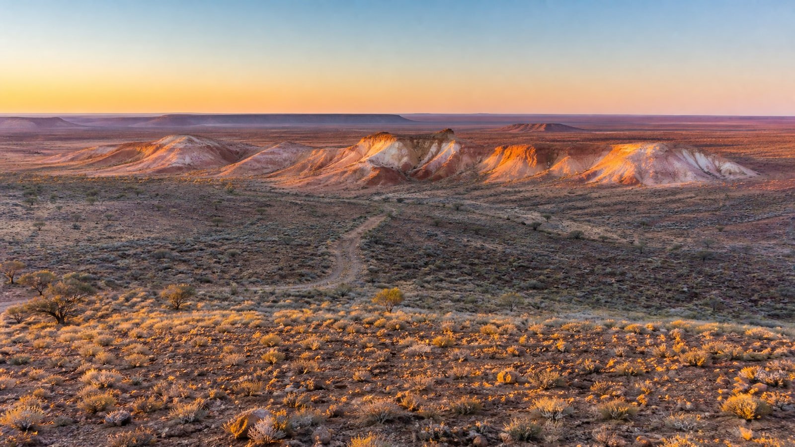 Sunrise over the colourful breakaway hills of Kanku-Breakaways Conservation Park near Coober Pedy, South Australia, with a faint dirt track crossing a stony gibber plain of saltbush and spinifex toward distant flat-topped mesas