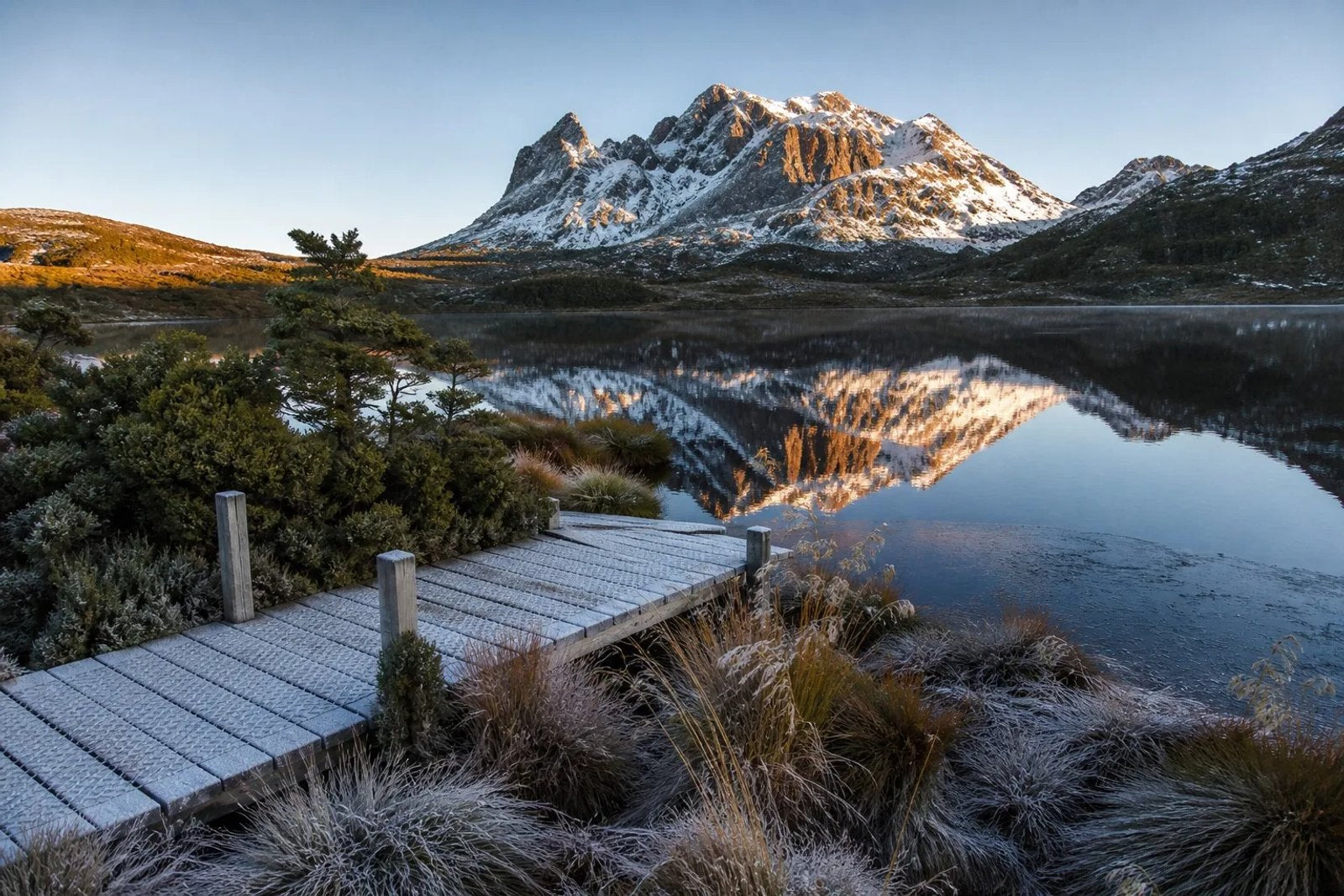 Snow-dusted Cradle Mountain peak reflected in the mirror-still water of Dove Lake at sunrise, a frosted timber boardwalk and buttongrass in the foreground, thin ice at the lake edge, Tasmania in late winter