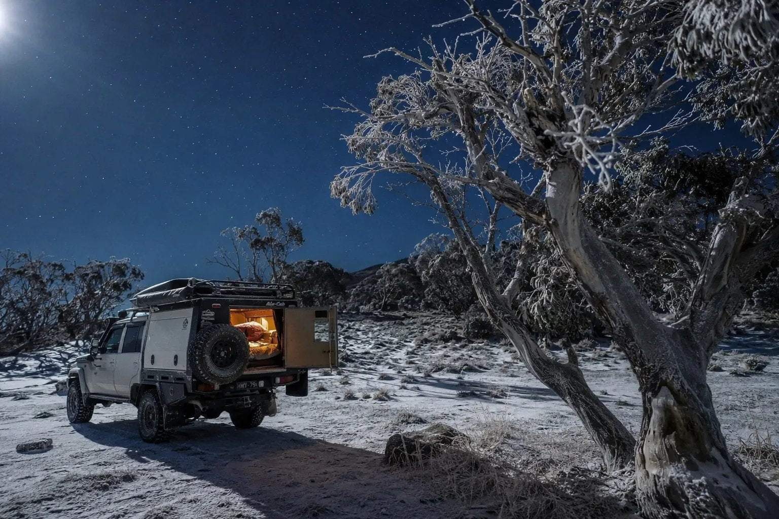 Rugged 4WD with a custom canopy parked at a remote alpine campsite in the Victorian High Country with a warm interior glow and snow-dusted trees under a starry winter sky.