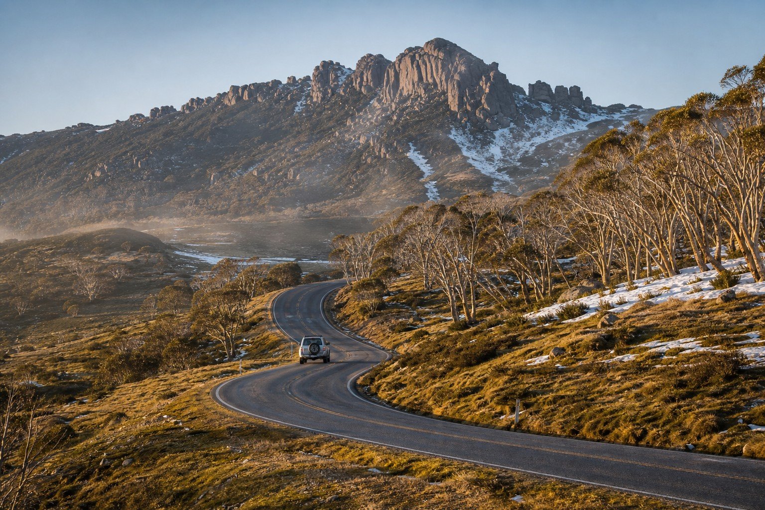 Winding bitumen of the Great Alpine Road climbing through snow gum woodlands towards the granite massif of Mount Buffalo in Victoria's High Country at morning, a white 4WD wagon on the road, last patches of snow on shaded slopes