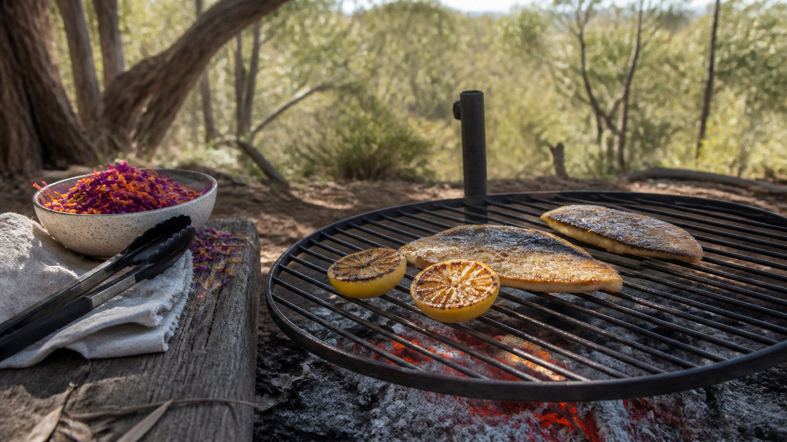 Skin-on snapper fillets grilling on a wire grill grate over glowing coals with charred lemon halves and a bowl of red cabbage slaw at an Australian bush camp
