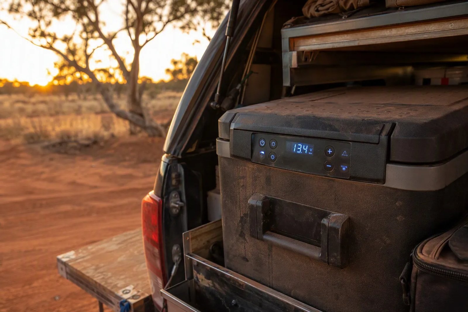 12V portable fridge in a 4WD canopy with its glowing digital control panel showing a battery voltage readout in a dusty Australian outback setting.