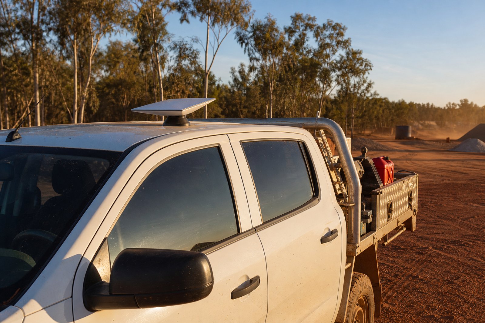 White dual-cab service ute parked on a red-dirt worksite with a compact Starlink Mini terminal mounted flat on the roof via a magnetic base, tool tray with diamond-plate storage box, eucalyptus and red dirt in golden light