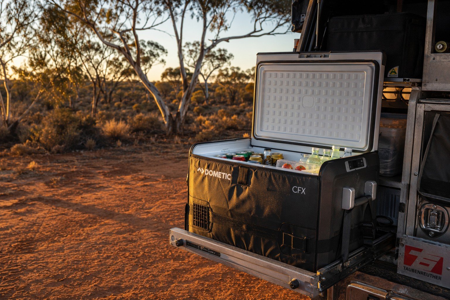 A 12V portable camping fridge with an insulation jacket, set up in the back of a 4WD vehicle during an outback sunset.
