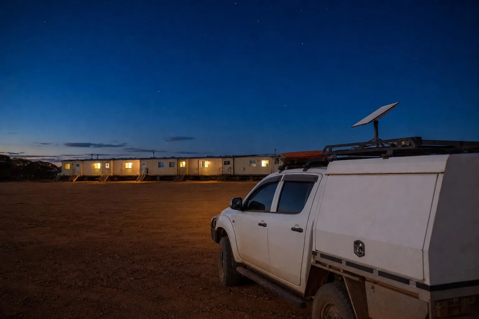 A Starlink Mini dish mounted on a service ute at a remote Australian mining camp during blue hour, with warm light glowing from modular accommodation dongas in the background.
