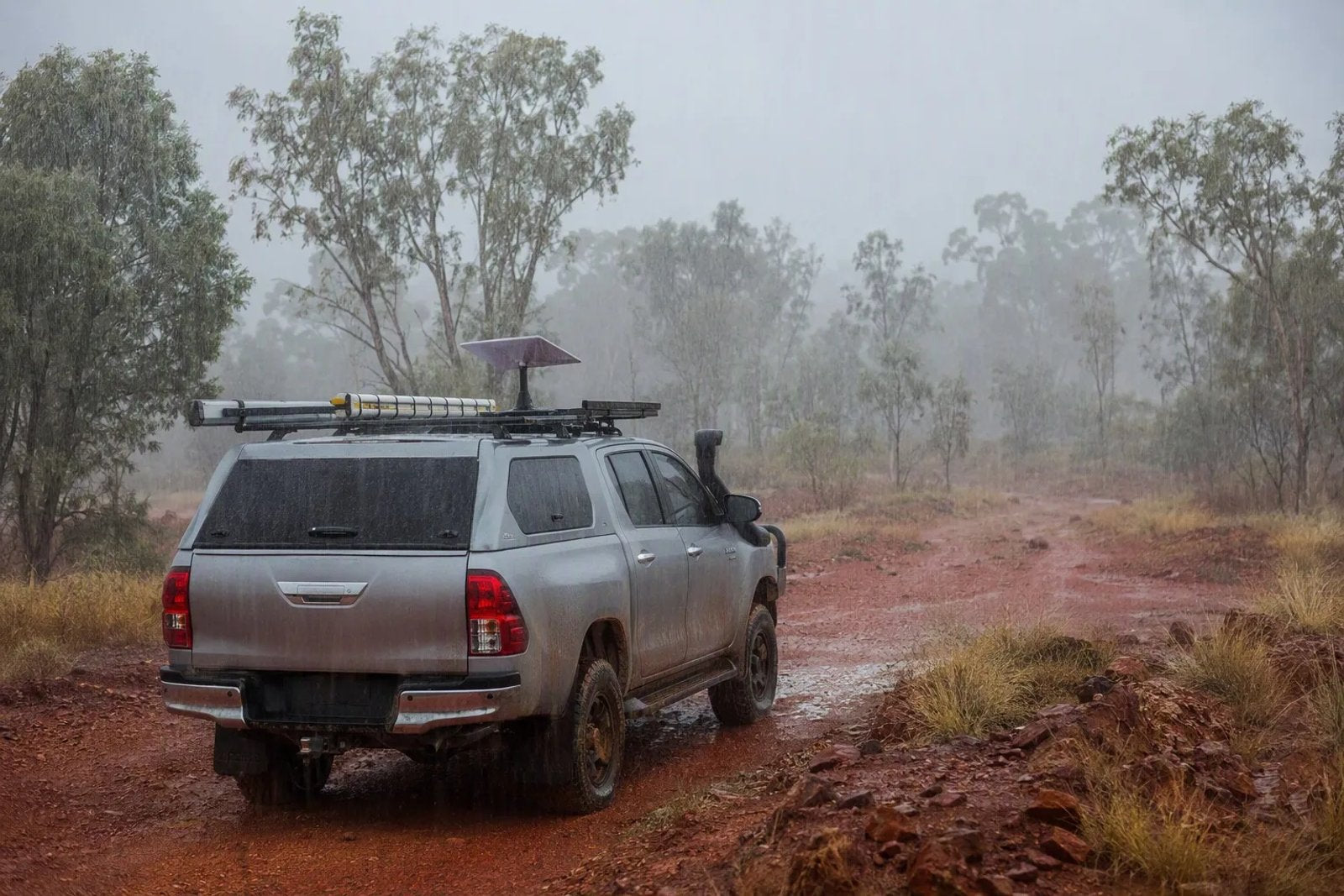 A Starlink Mini dish mounted on the roof rack of a rugged 4WD ute during a heavy tropical rainstorm in the Australian bush.
