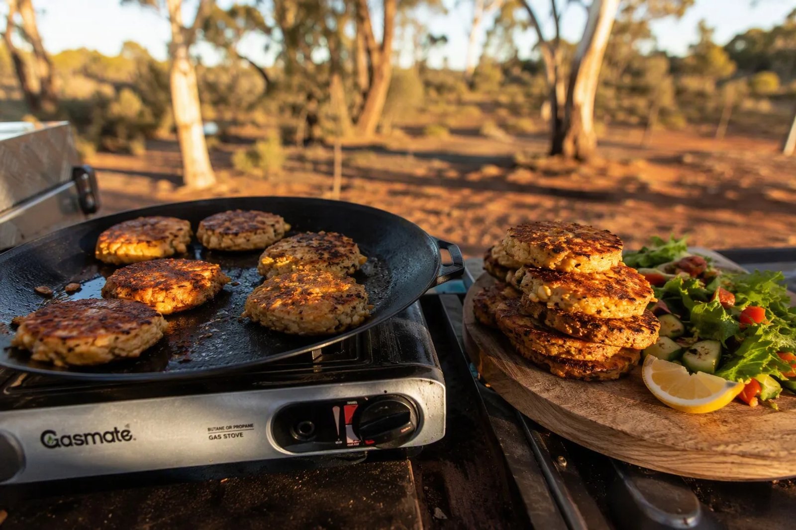 Golden-brown crispy tuna and sweet corn patties sizzling on a cast-iron hotplate over a portable gas stove at an Australian bush camp, with a stack on a wooden board with salad and lemon at golden hour.