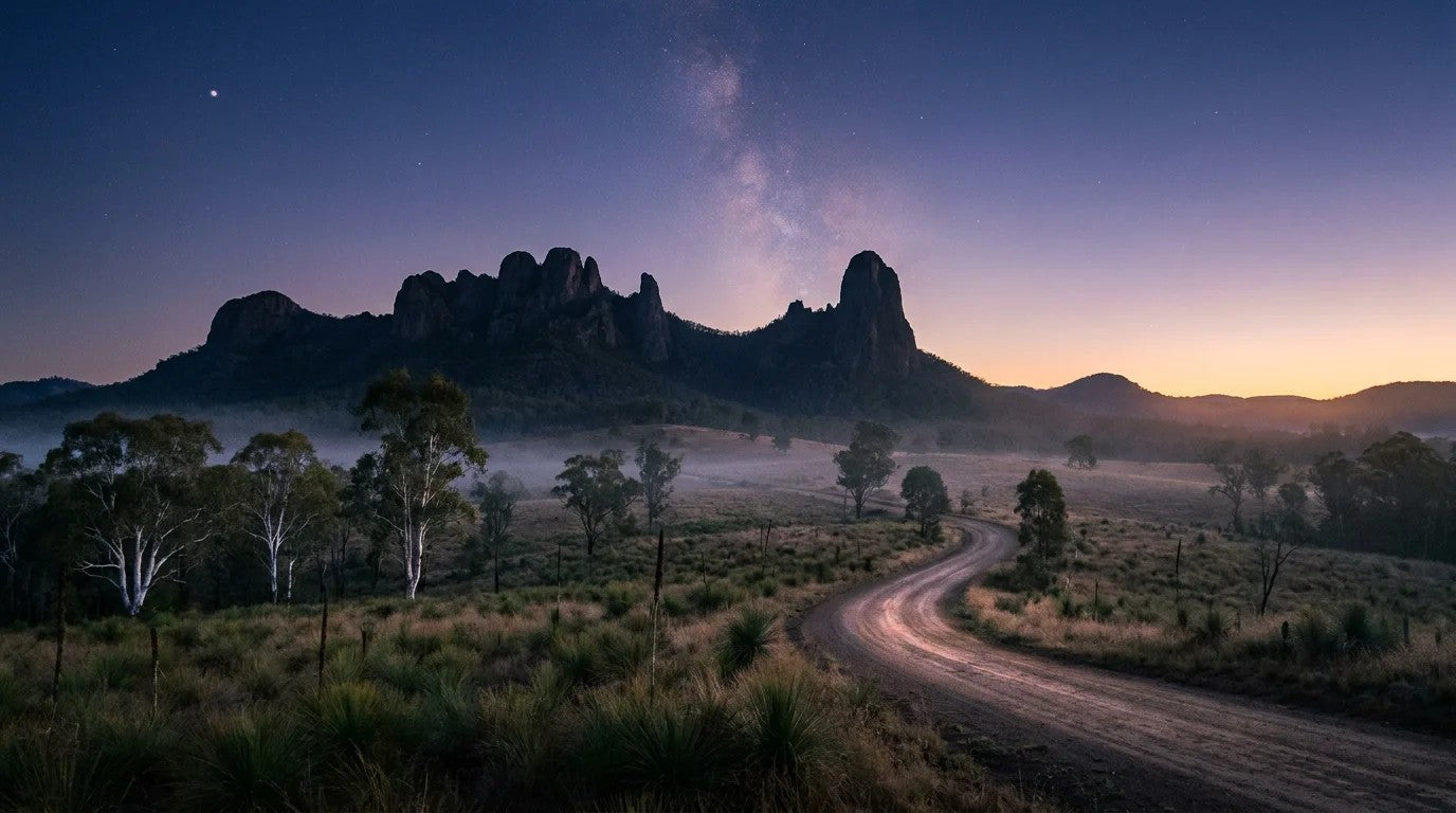 Pre-dawn blue hour at Warrumbungle National Park NSW, the silhouette of the Breadknife and Belougery Spire against a deep cobalt sky with the Milky Way fading, mist over native grasses, white-trunked gums and a dirt road curving in the foregr...