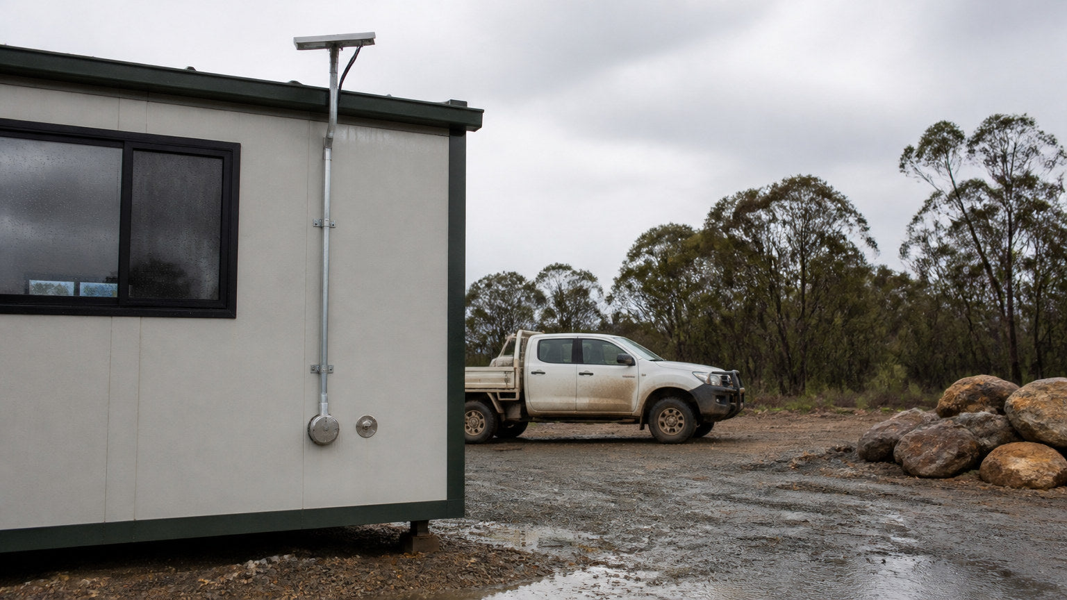 Satellite terminal on the roof of a demountable site office with conduit running down a galvanised pole to a sealed cable gland on the wall, wet gravel and a dusty ute in the background