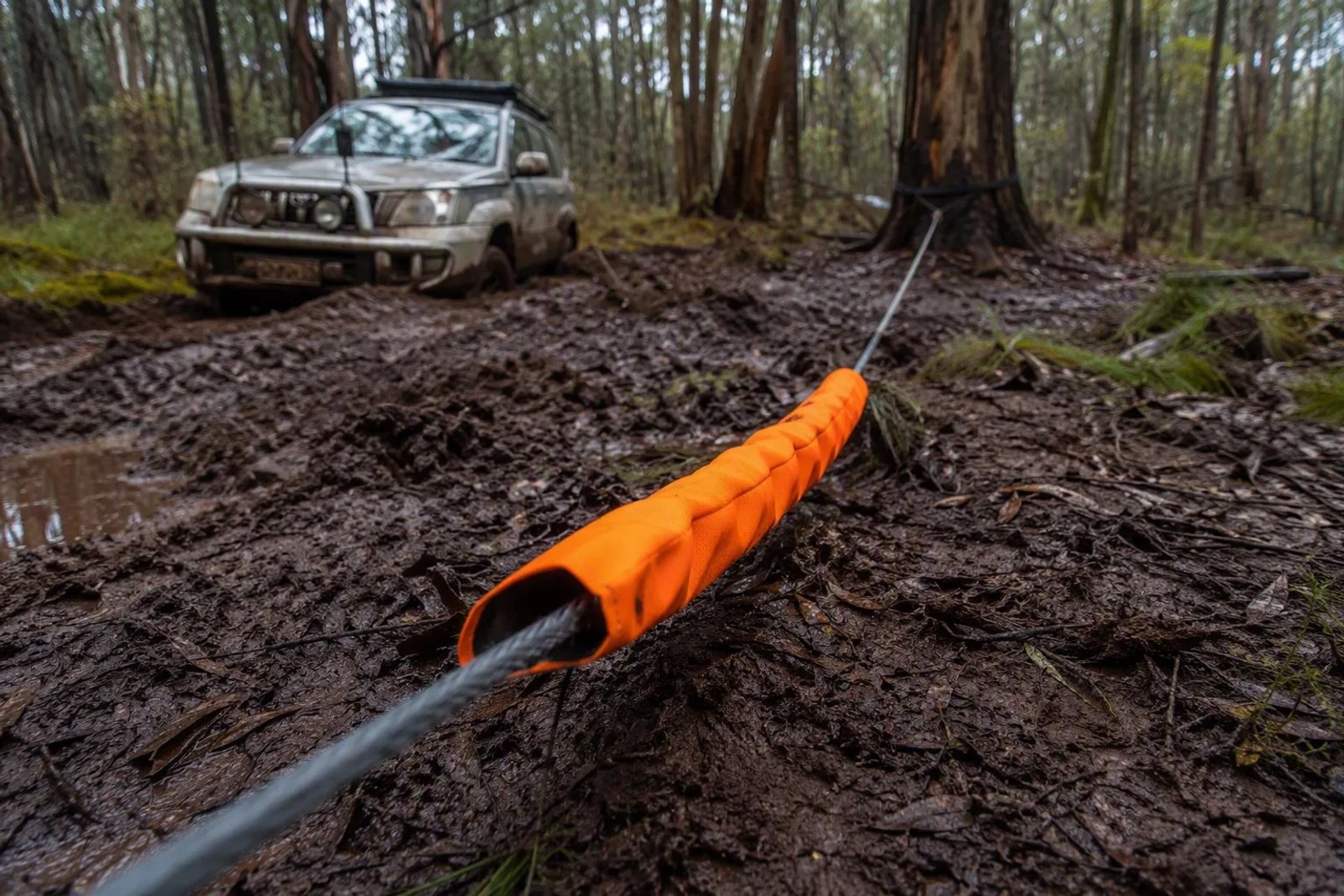 4WD recovery scene on a muddy Australian winter track with a bright orange winch damper draped over the line.